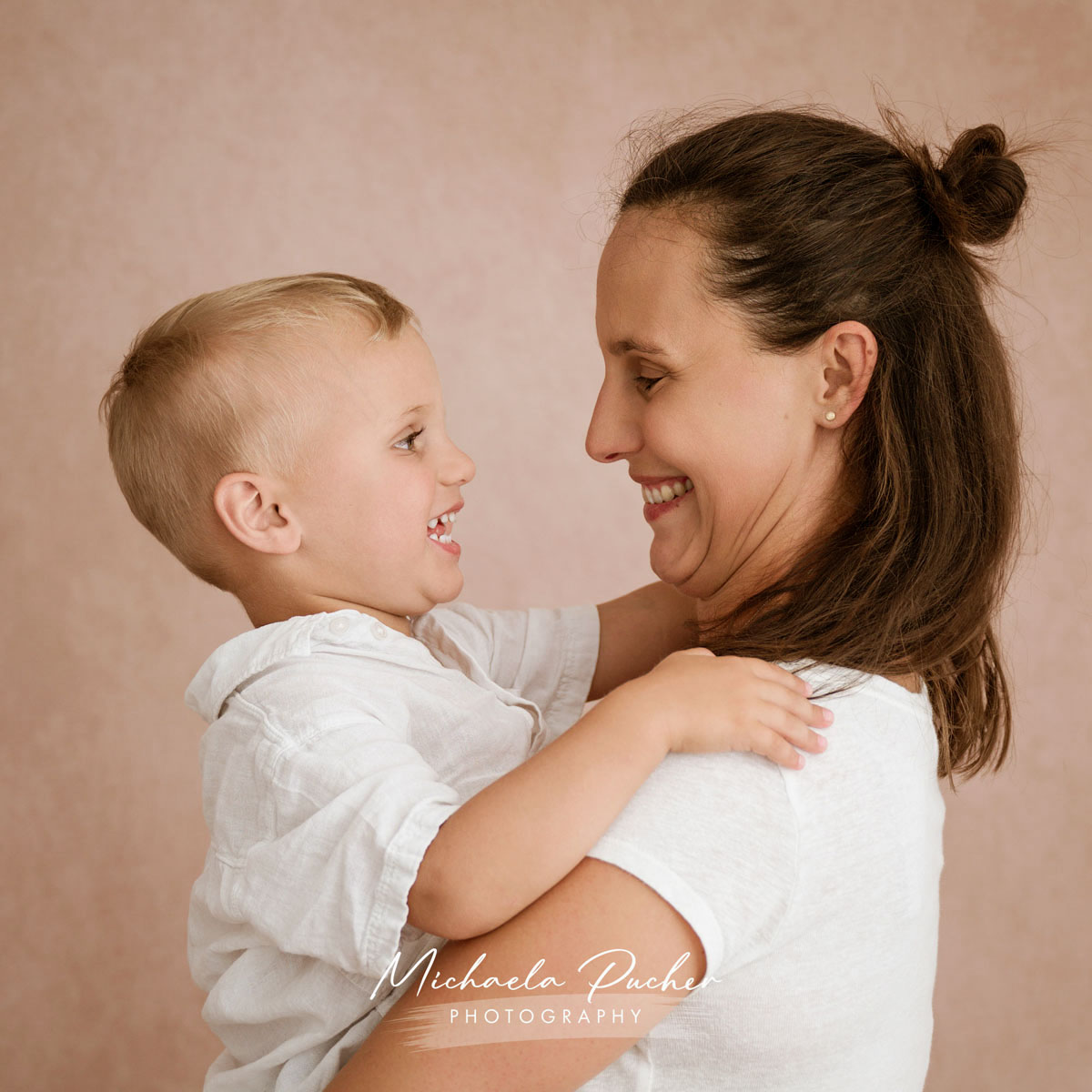 Mutter hält ihr lachendes Kind im Arm, beide blicken sich fröhlich an vor einem zarten rosigen Hintergrund.